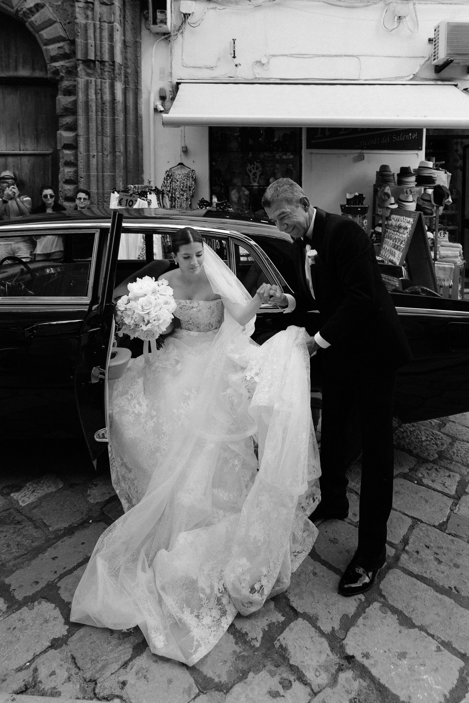 Black and white photo of a formal wedding moment with an elegantly dressed couple exiting near a vintage car on cobblestones.