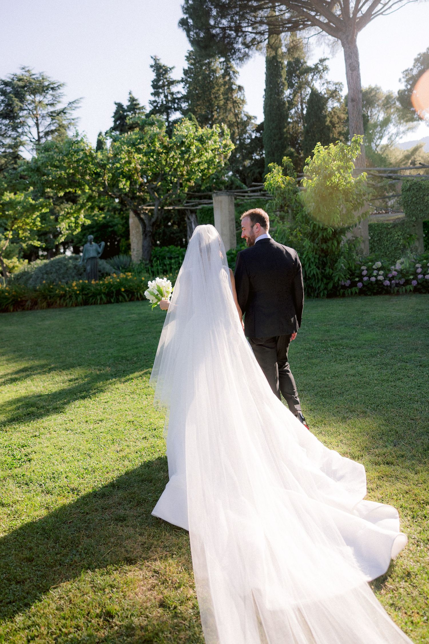 Wedding photo taken in a lush garden setting with a long flowing white veil trailing behind the couple on a sunny day.