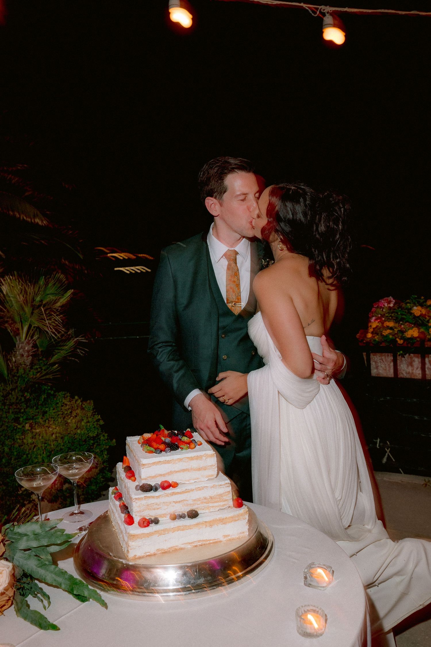 A wedding couple shares a kiss next to their white three-tiered wedding cake decorated with fresh strawberries and lit candles.