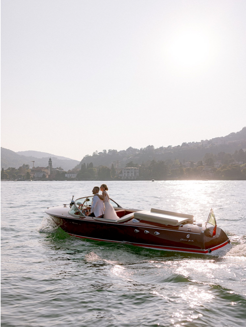 Couple riding a classic wooden speedboat on a sunlit lake with scenic hills and a village in the background.