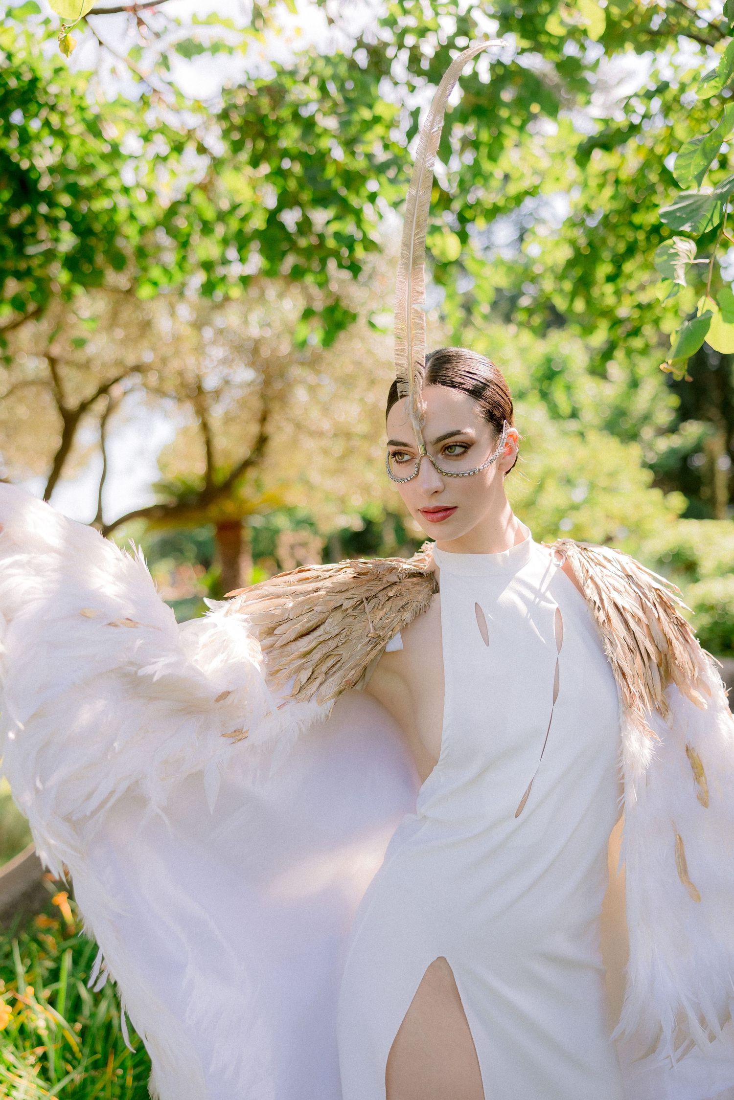 Woman in elegant white gown with feather wing accents poses outdoors in a sunlit garden with green foliage.