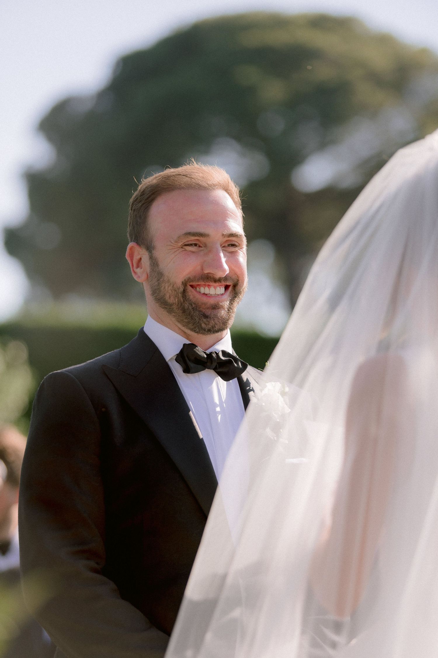 A groom in a black tuxedo and bowtie smiles joyfully during an outdoor wedding ceremony.