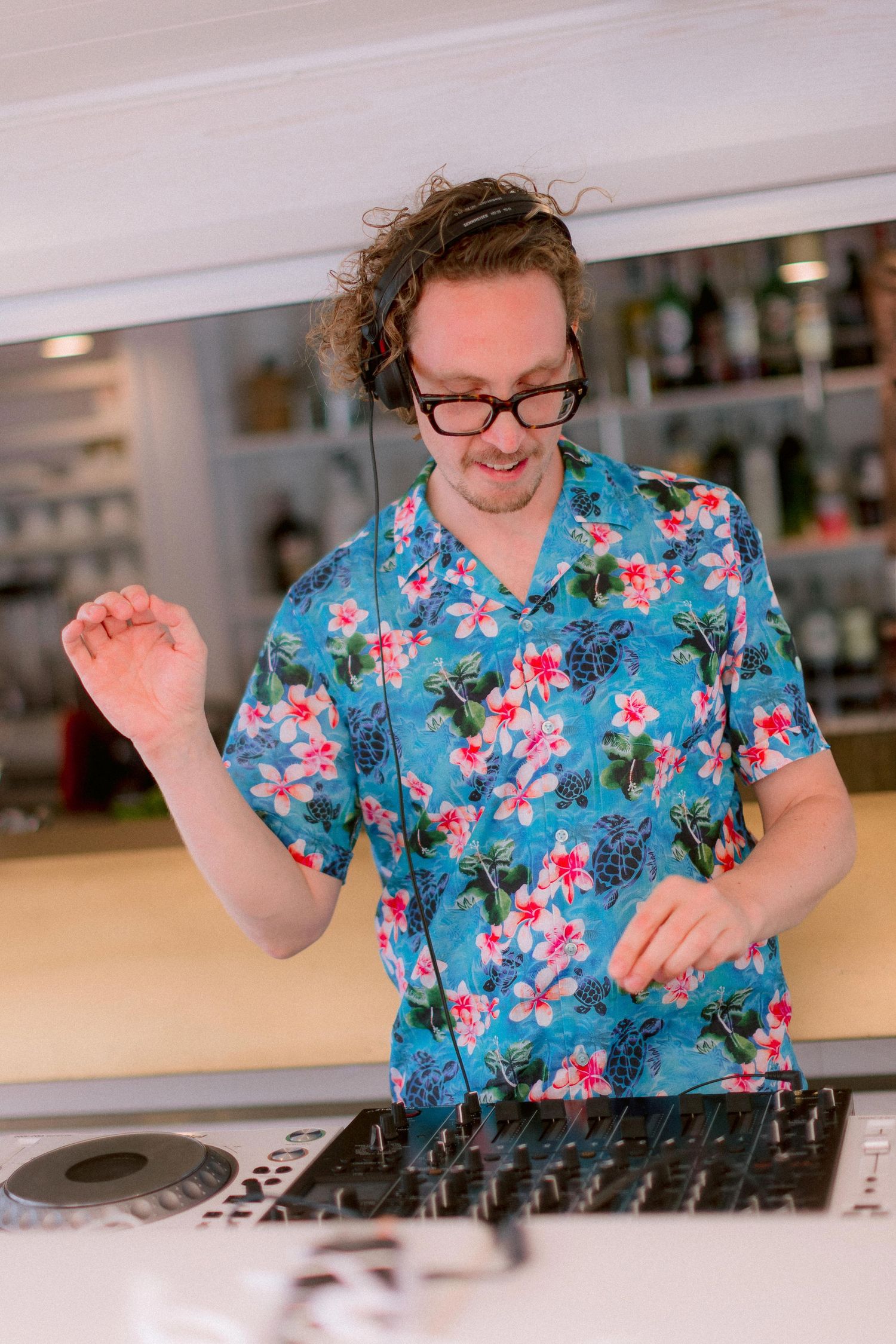 DJ in a vibrant blue floral Hawaiian shirt performs at a mixing console with enthusiastic movement.