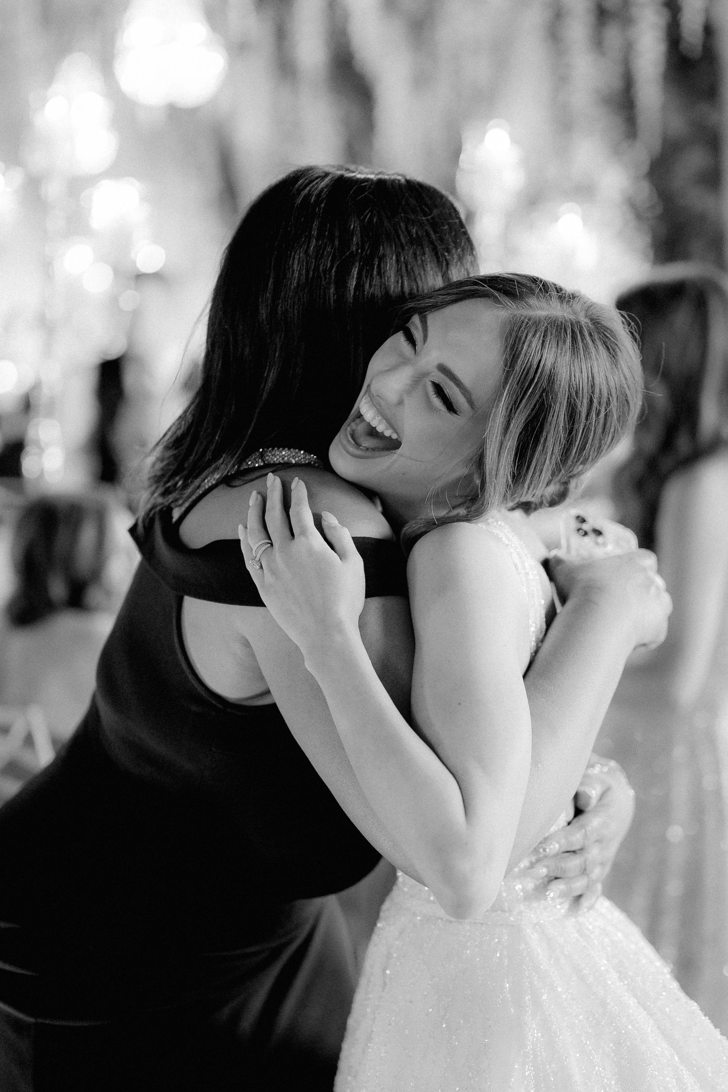 Black and white photo of emotional embrace between two women, one in a dark dress and one in white bridal gown.