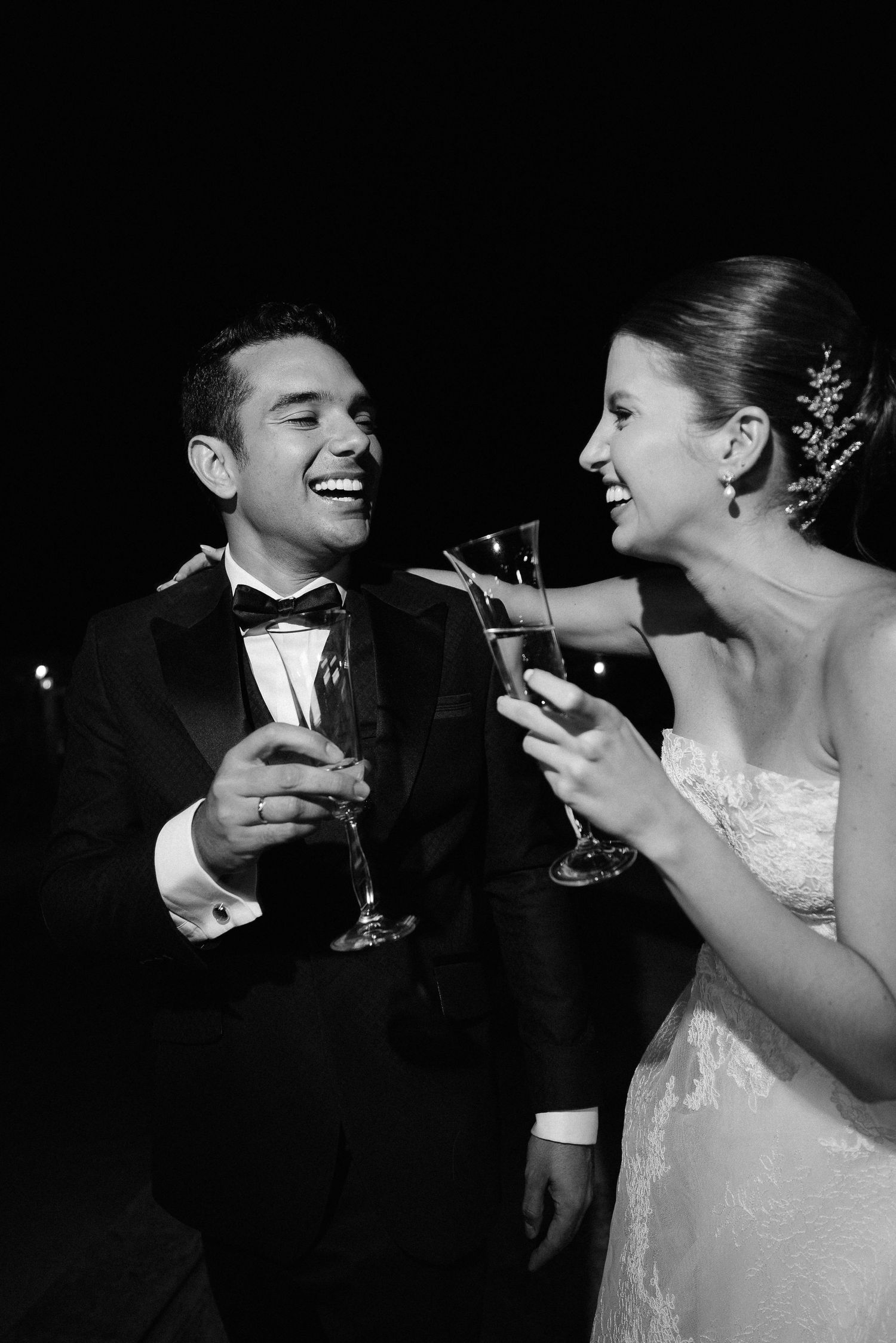 Couple in formal attire laughing while holding champagne glasses at elegant black and white celebration.