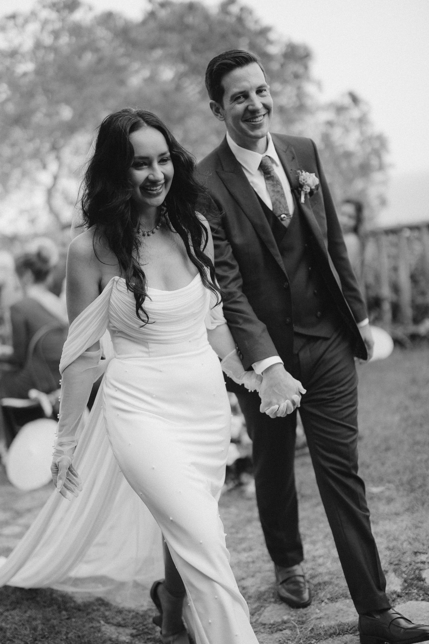 A joyful bride in a flowing white dress and groom in dark suit walking together outdoors in black and white photography.