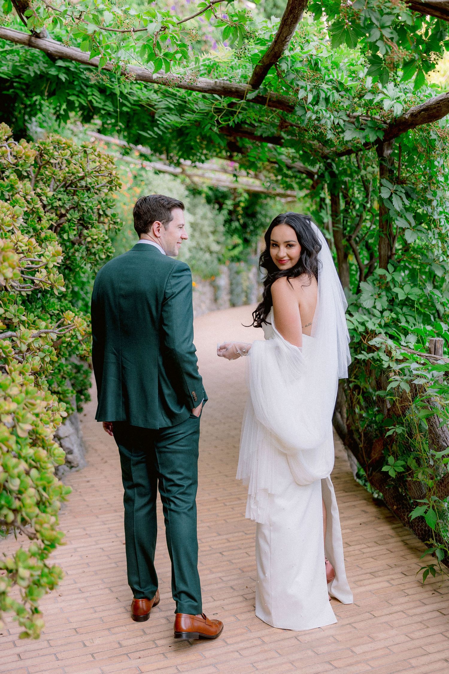 A couple in wedding attire walking along a garden path under a canopy of lush green vines and foliage.