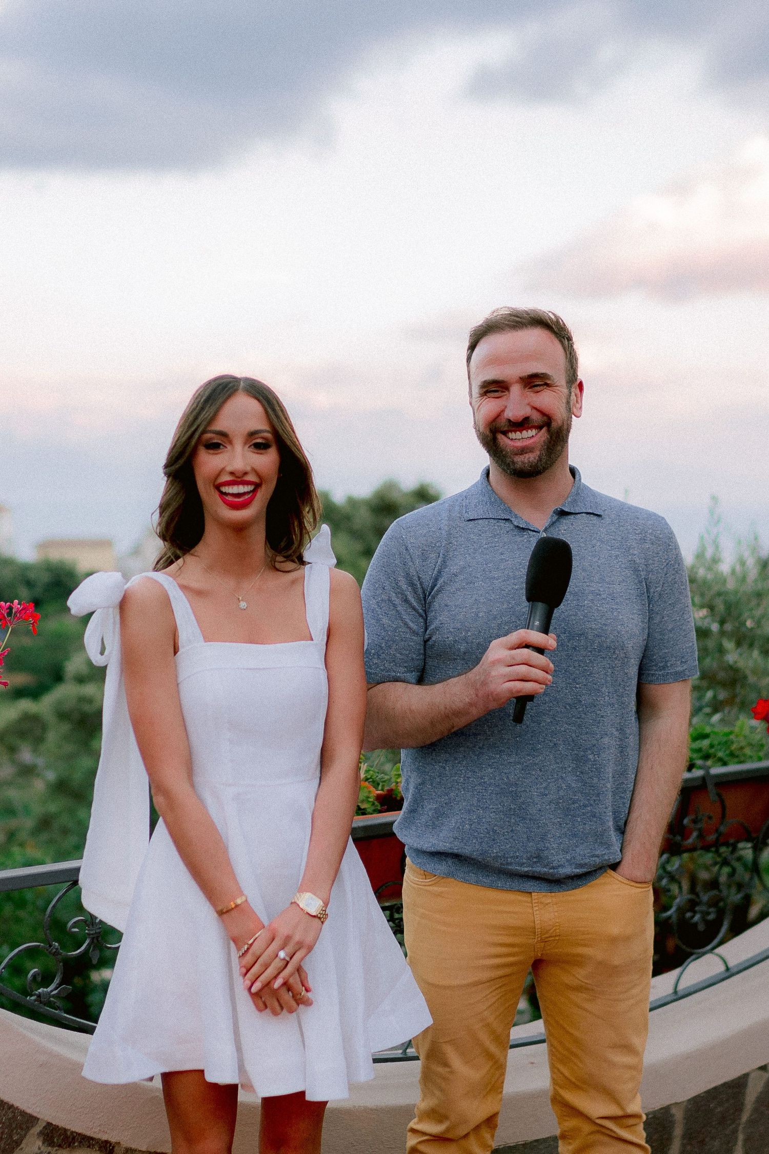 Two people standing outdoors, one in a white dress and one in grey polo holding microphone, against cloudy sky at dusk.