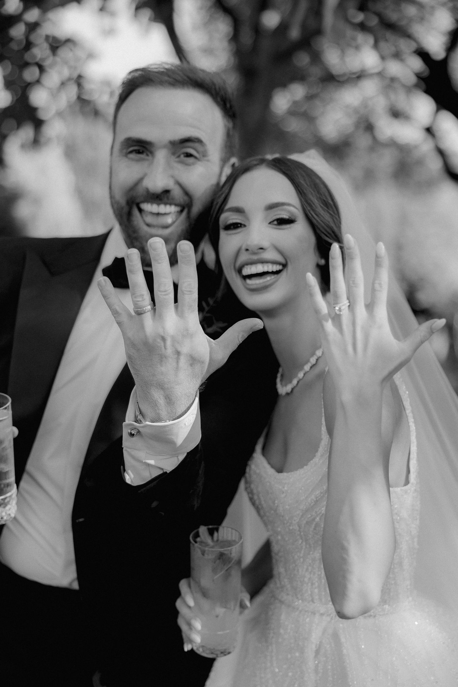 Black and white photo of newlyweds showing off their wedding rings with joyful smiles outdoors.