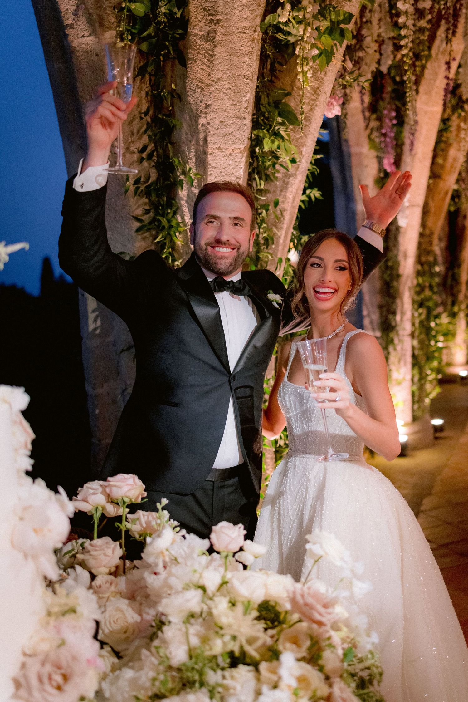A couple dressed in wedding attire raise their hands joyfully at an evening reception with illuminated trees and floral decor.