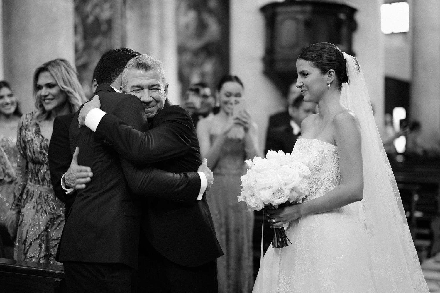 A black and white wedding ceremony moment captured inside a church with two people embracing while a bride holds white flowers.