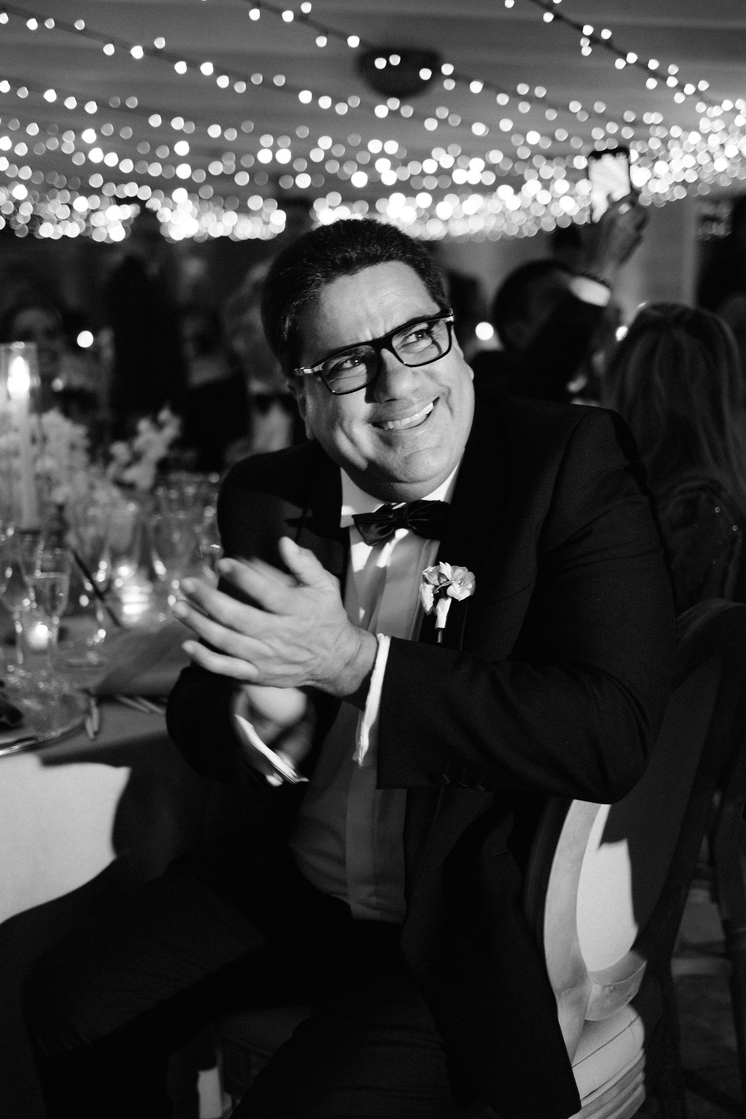 A guest in a black tuxedo and glasses laughs joyfully at an evening celebration with twinkling lights overhead.
