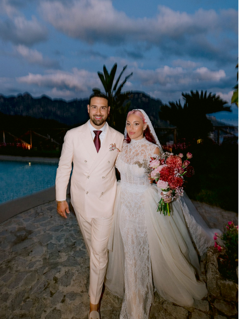 Bride in lace gown holding pink bouquet stands with groom in cream suit by pool at outdoor wedding ceremony.