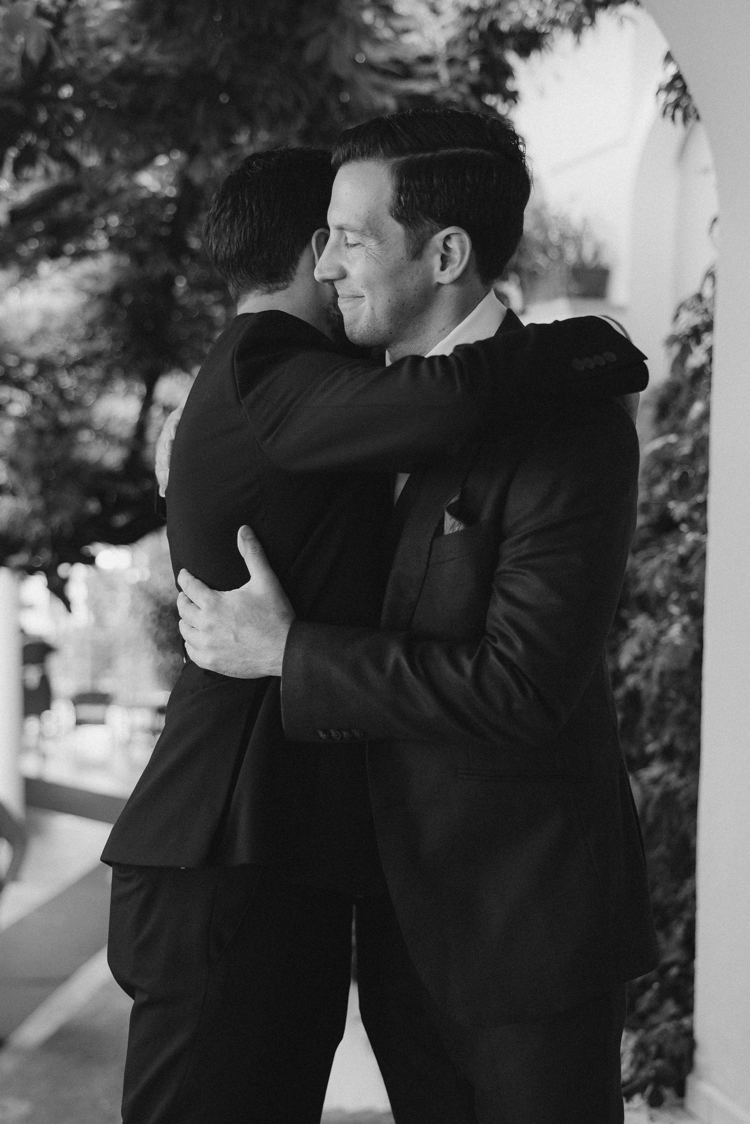 Two people in black suits share an emotional embrace in a black and white outdoor setting under an archway.