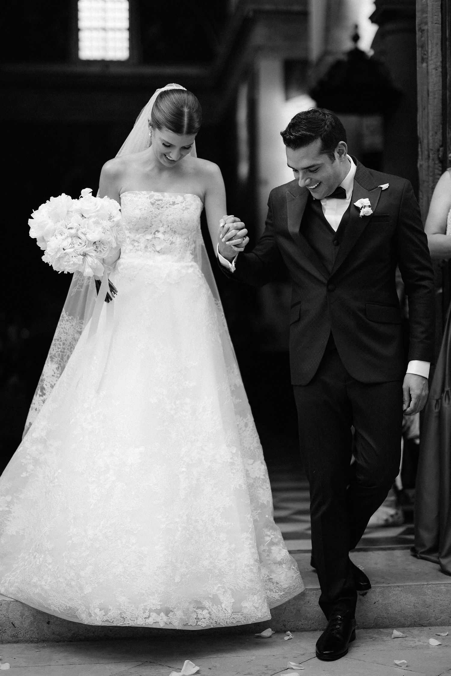 A bride in a flowing lace gown holds a white bouquet as she walks with her groom in a classic black tuxedo.