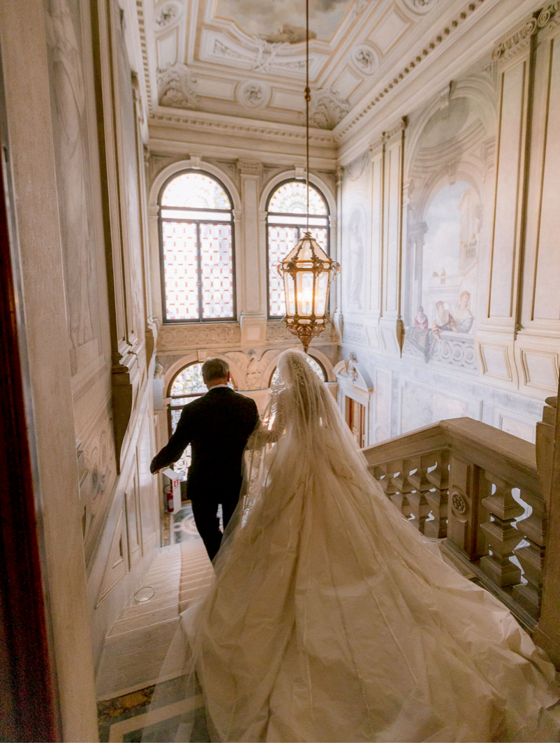 Bride in elegant gown with long veil walks with groom down ornate marble staircase in grand historic venue.