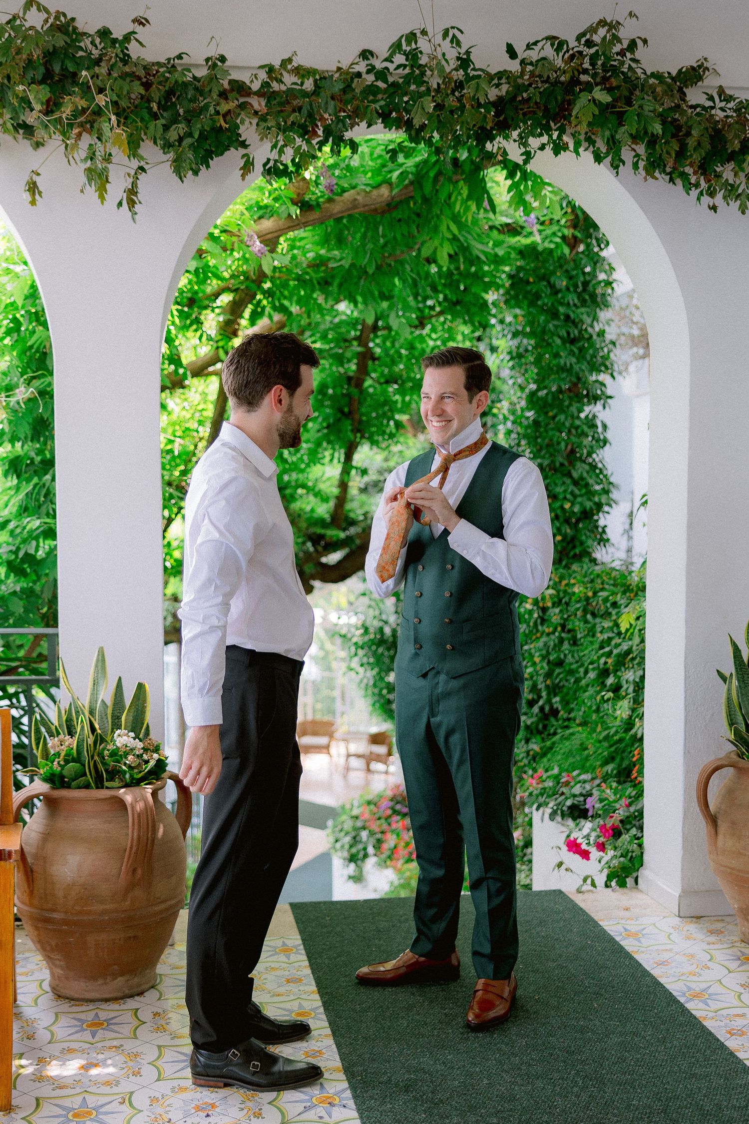 Two people share a conversation under an arched walkway covered with lush green vines and potted plants.