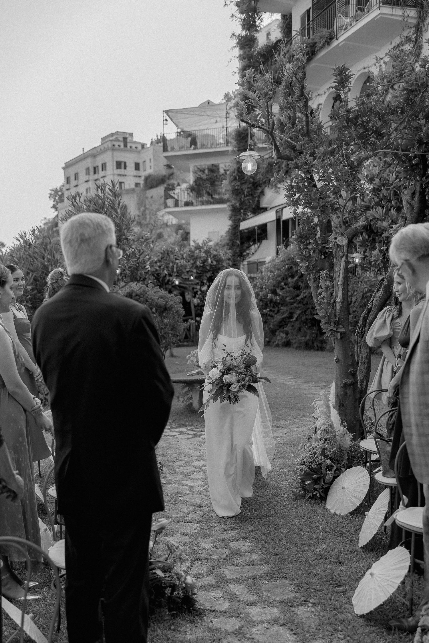 Black and white wedding scene in Positano showing garden path with guests and Mediterranean hillside architecture.