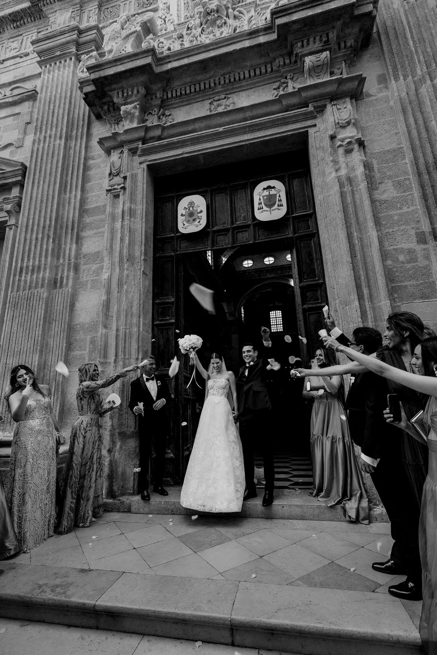 A wedding party exits a grand stone church entrance while guests line the steps throwing confetti in black and white.