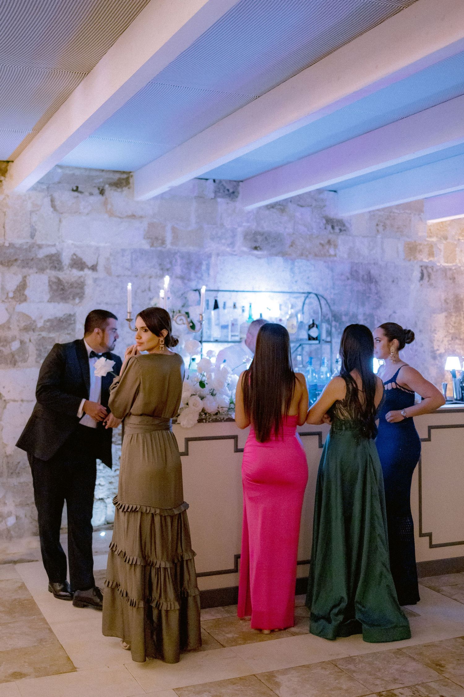 A group of formally dressed guests gather around a sleek bar area with stone walls illuminated by blue lighting.
