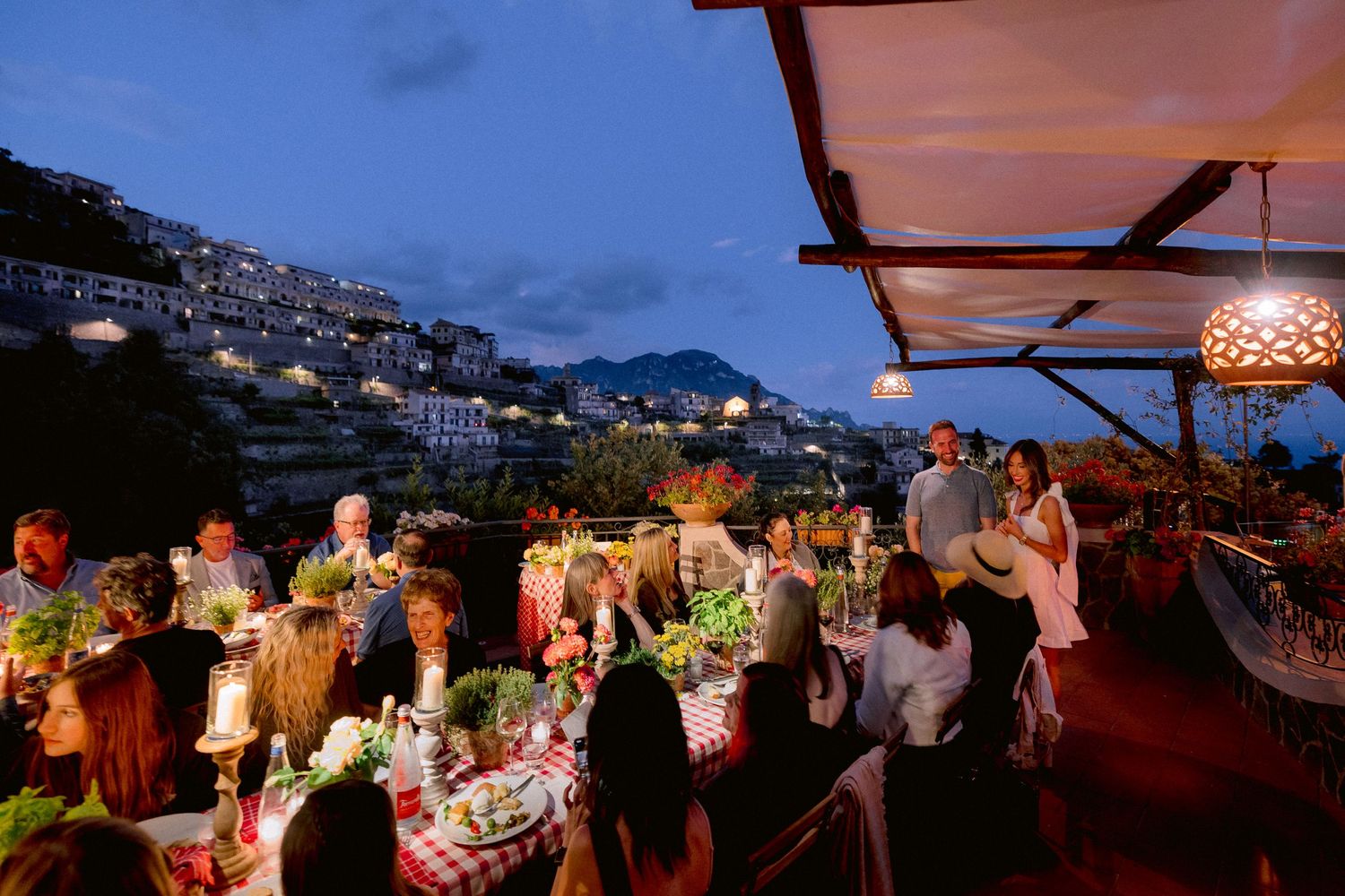 An elegant outdoor dinner party on a rooftop terrace overlooking a hillside town at dusk with glowing lanterns.