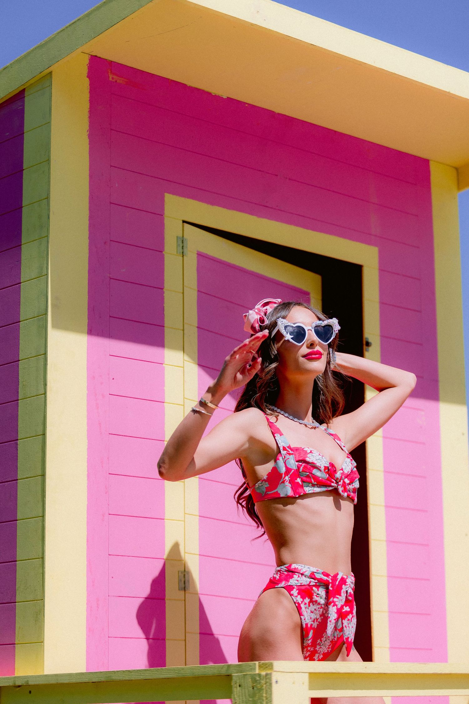 Colorful beach cabana with bright pink wall and yellow trim featuring someone in a red floral swimsuit and retro sunglasses.