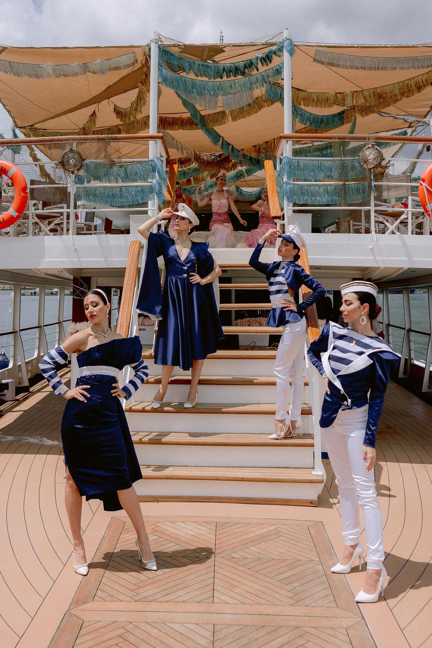 Dancers in navy and white nautical costumes pose on wooden stairs of a cruise ship deck under decorative canopies.