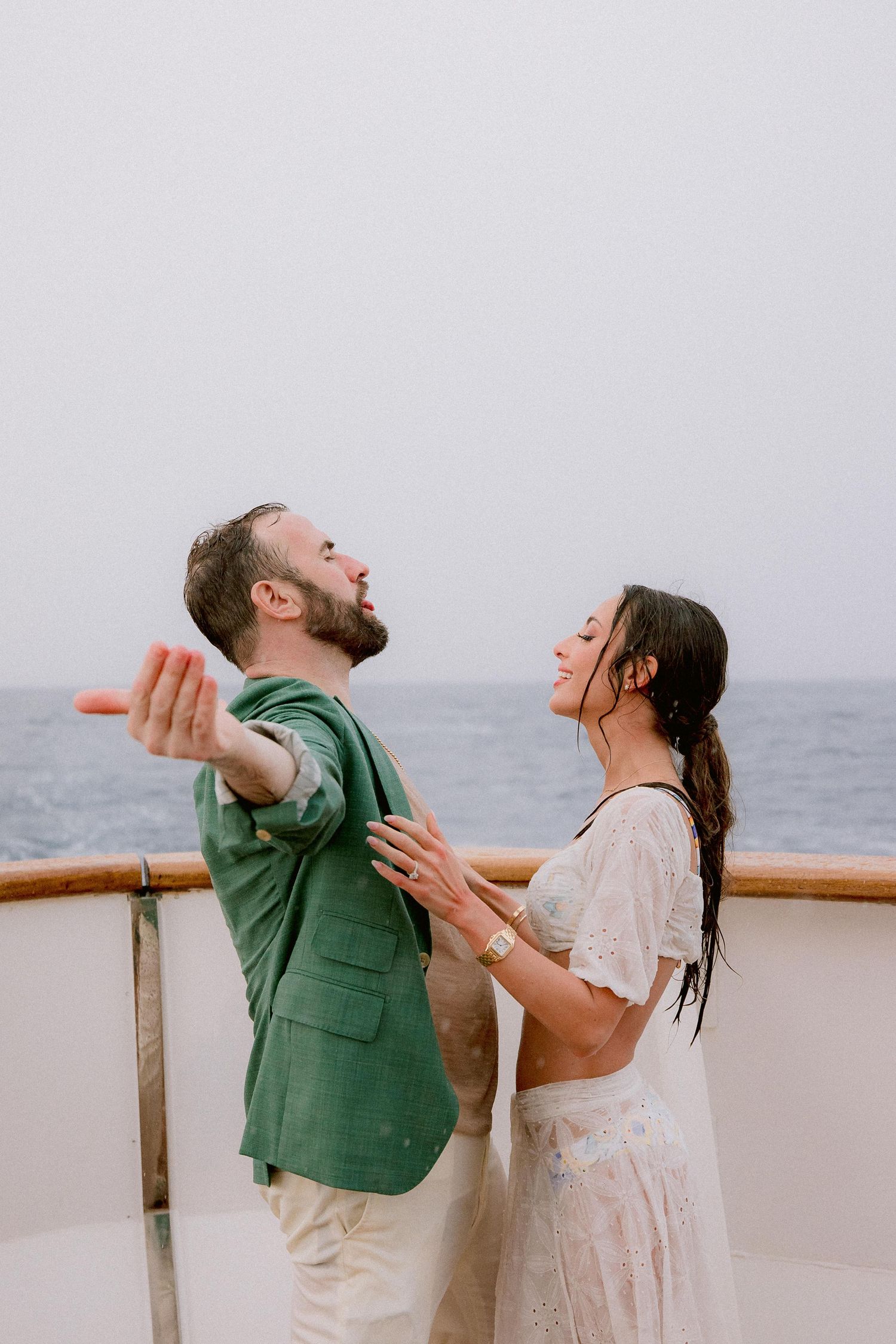 A joyful couple laughs together on a boat deck with the ocean and overcast sky in the background.