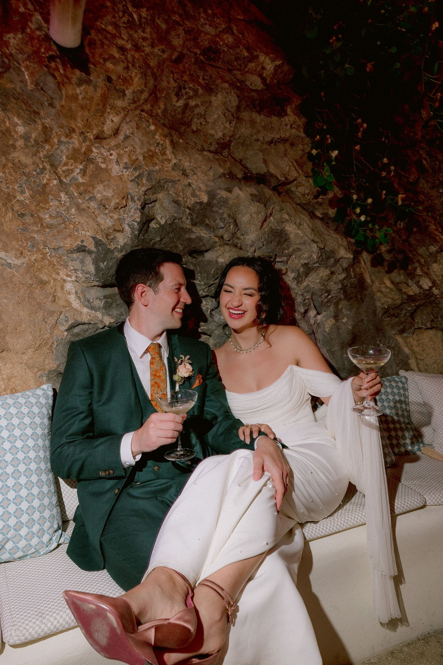 A couple in wedding attire shares a joyful moment on a sofa in a rustic stone-walled setting while holding drinks.
