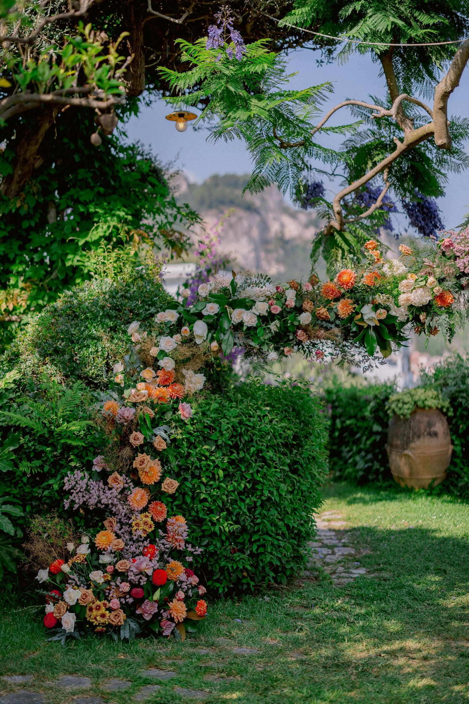 A romantic garden arch decorated with peach roses and purple flowers frames a winding path through lush greenery.