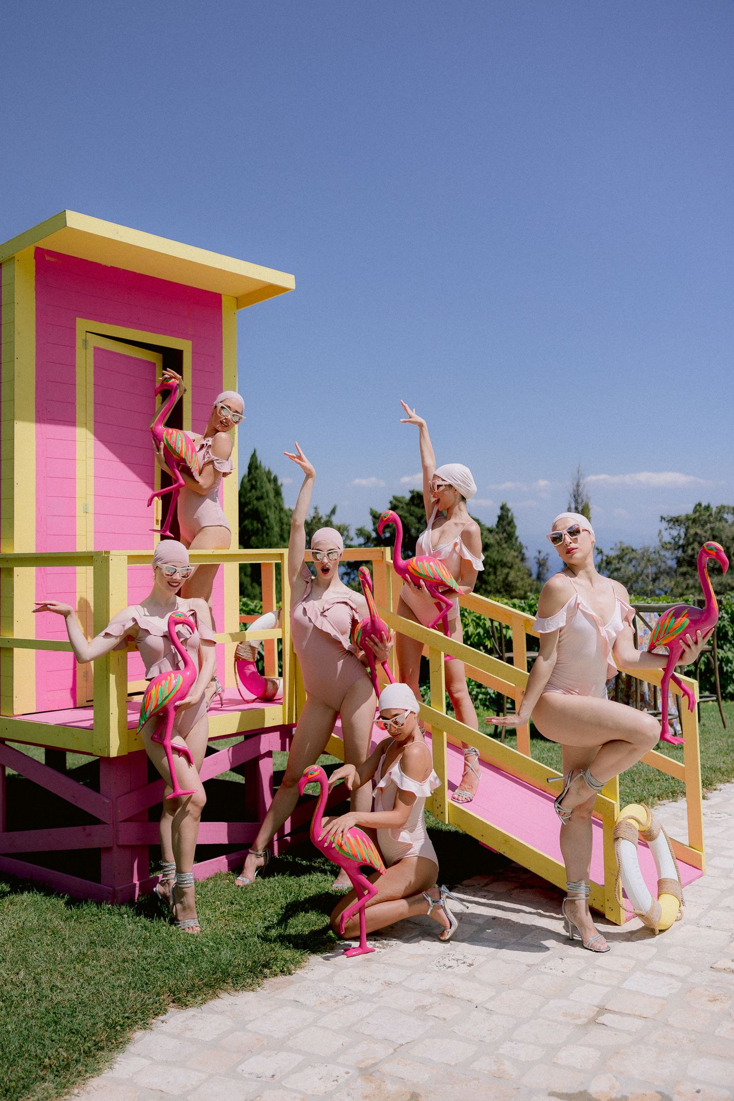 People in pink swimsuits pose playfully around a colorful yellow and pink lifeguard tower at a retro-styled photoshoot.