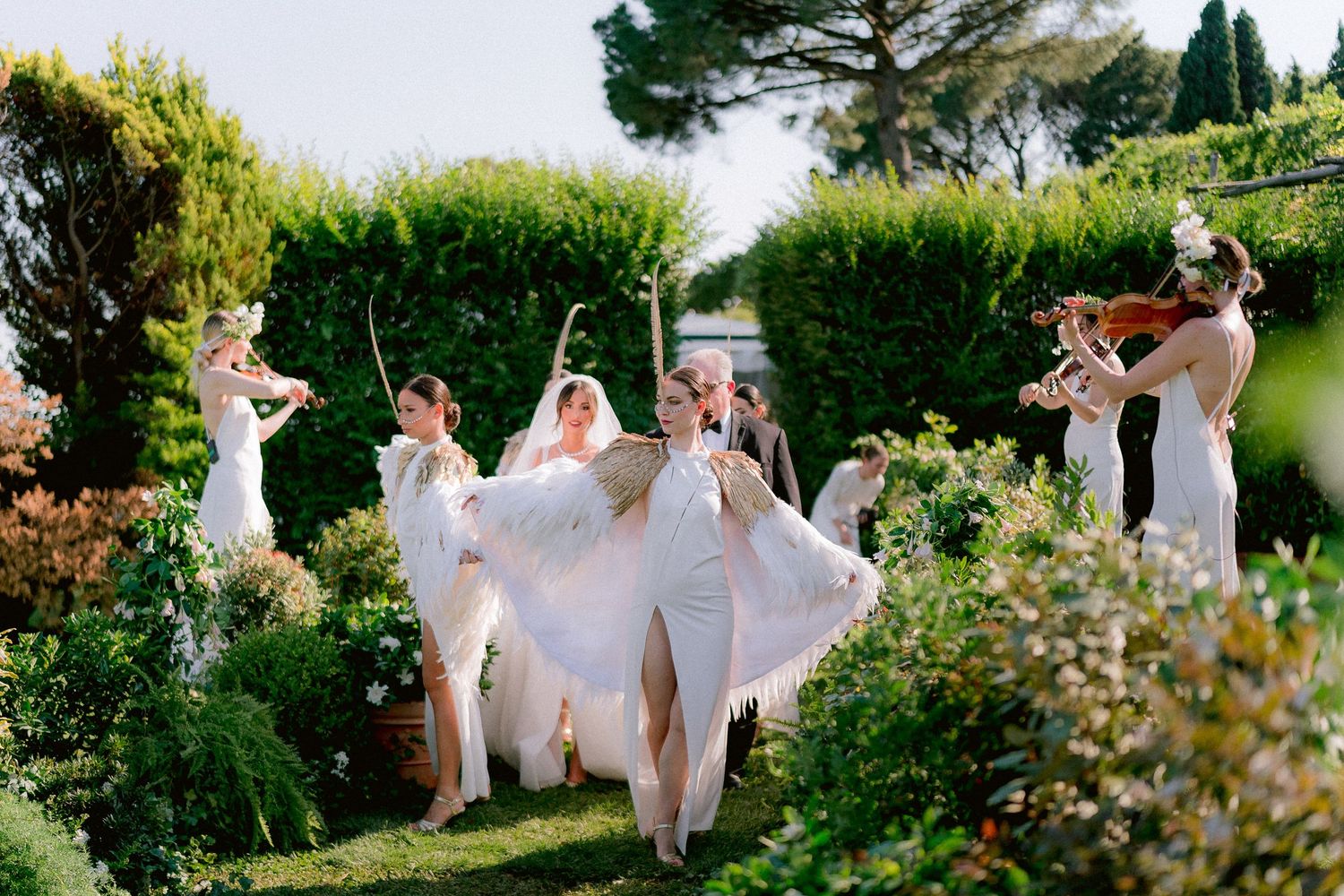 Group in white dresses and suits dance in a garden surrounded by blooming flowers and greenery during a celebration.