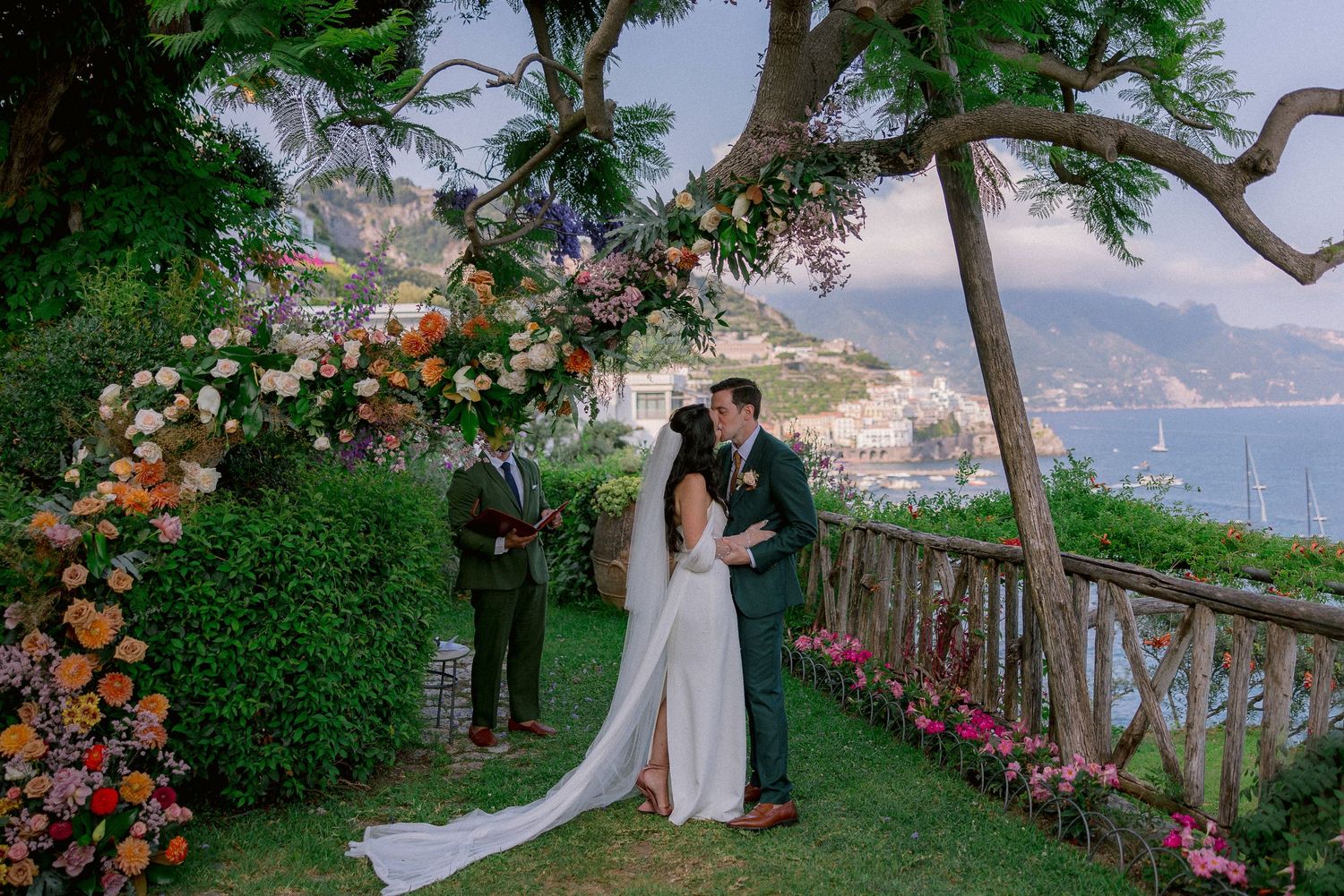 Wedding ceremony takes place under floral arch at scenic coastal overlook with ocean and mountains in background.