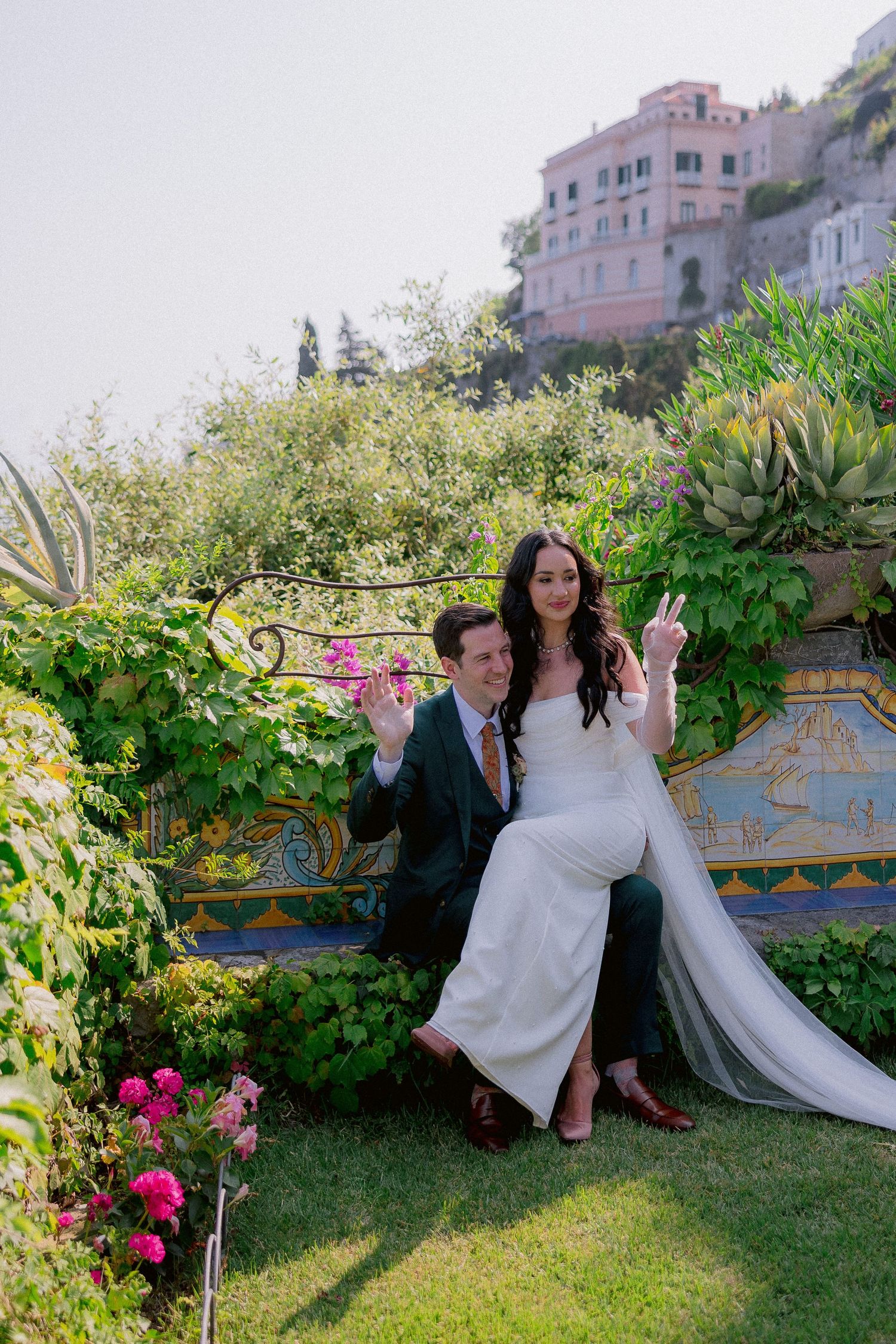 Wedding couple sits on decorative tiled bench in lush Mediterranean garden with pink villa in background.