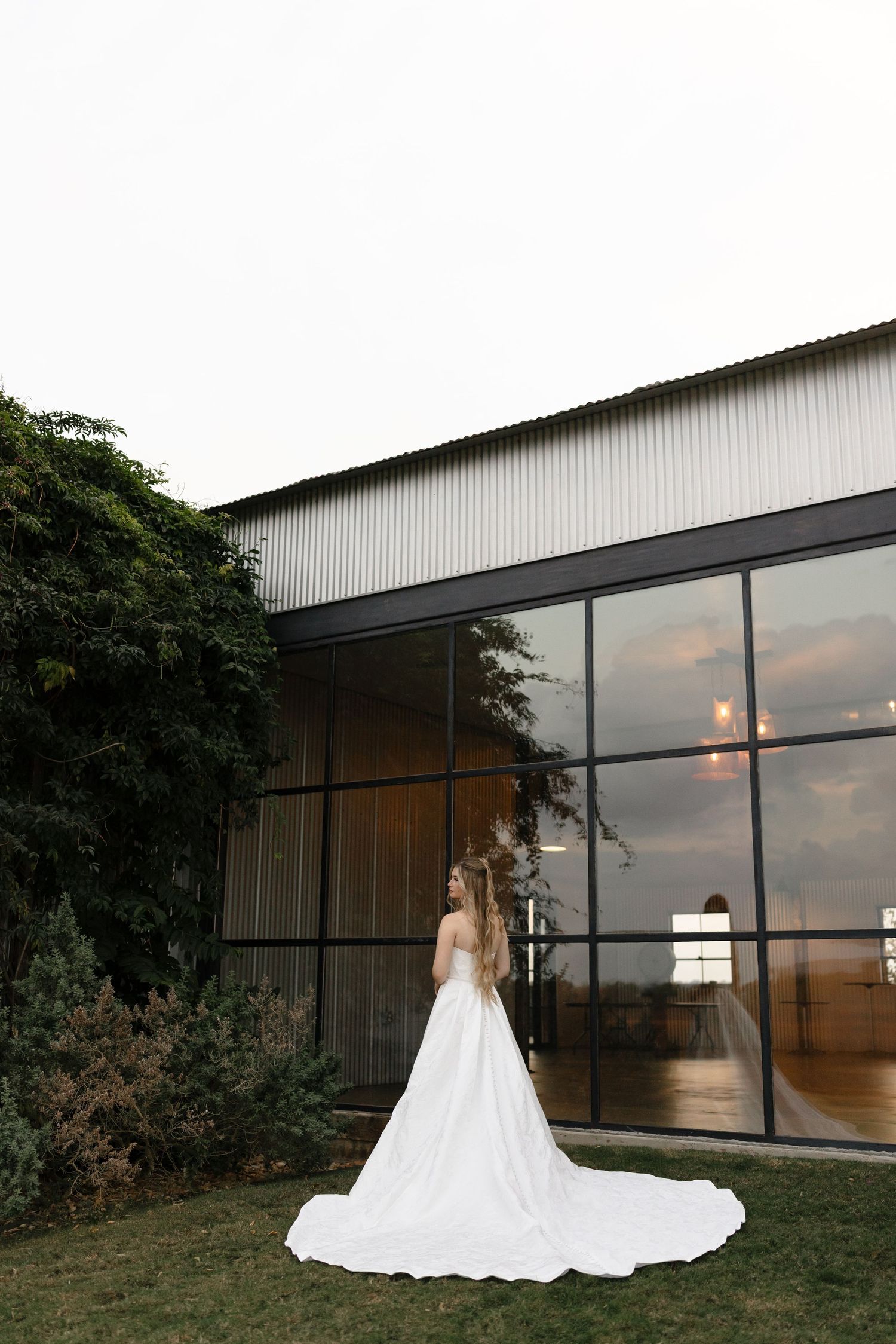 A bride in a flowing white wedding dress stands silhouetted against a modern building at sunset.