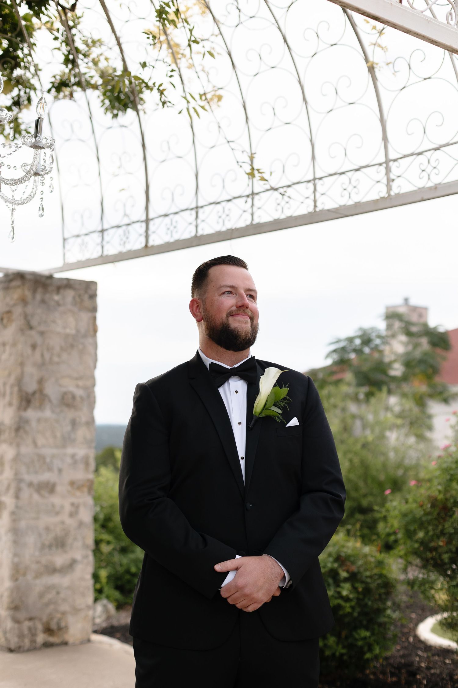Groom in black tuxedo smiles and poses outdoors against stone pillars and greenery during wedding photoshoot.