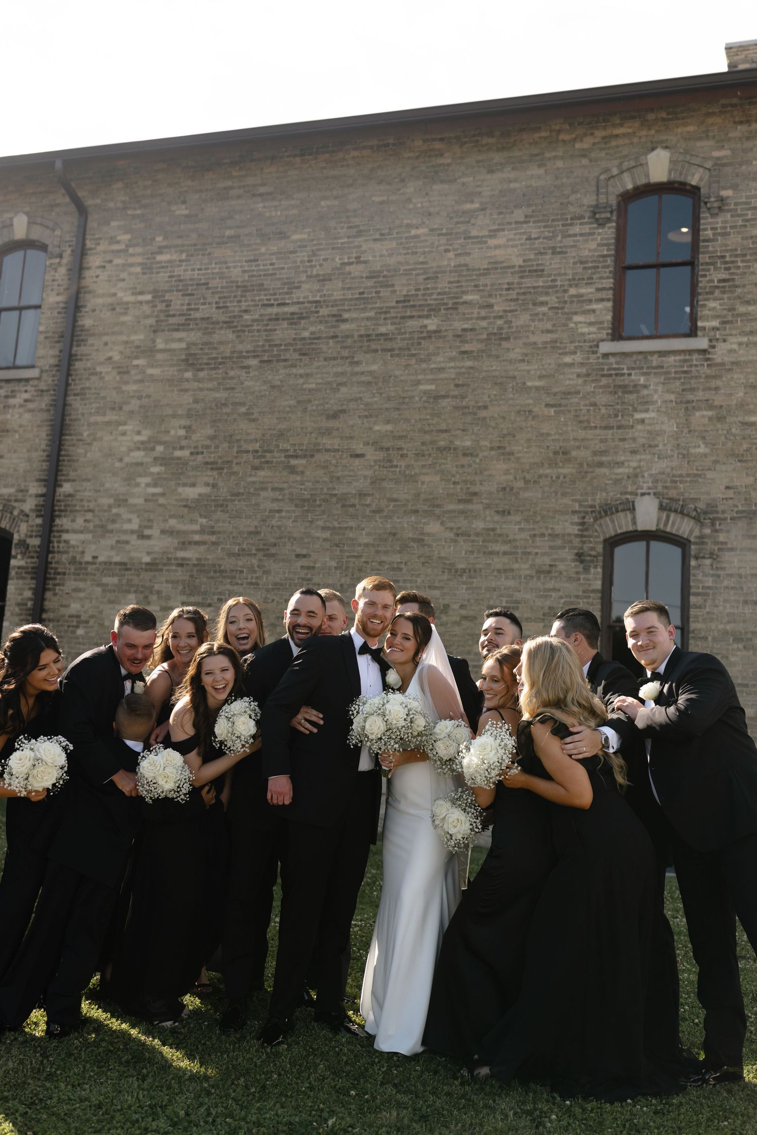 Wedding party wearing black and white attire poses together against a brick wall outdoors.