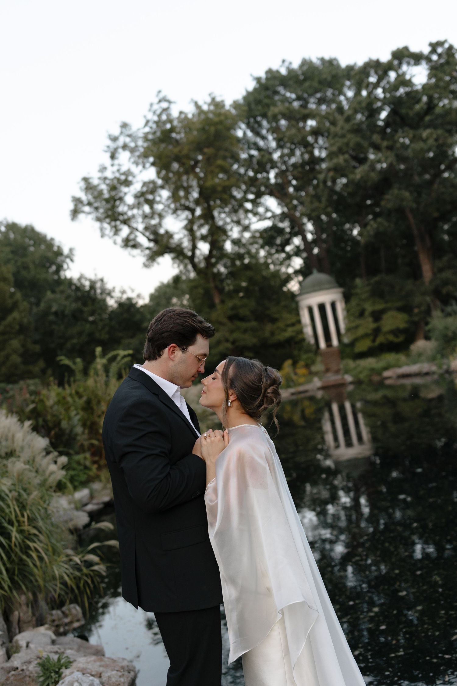 A couple shares an intimate moment by a reflective pond surrounded by lush greenery at dusk.