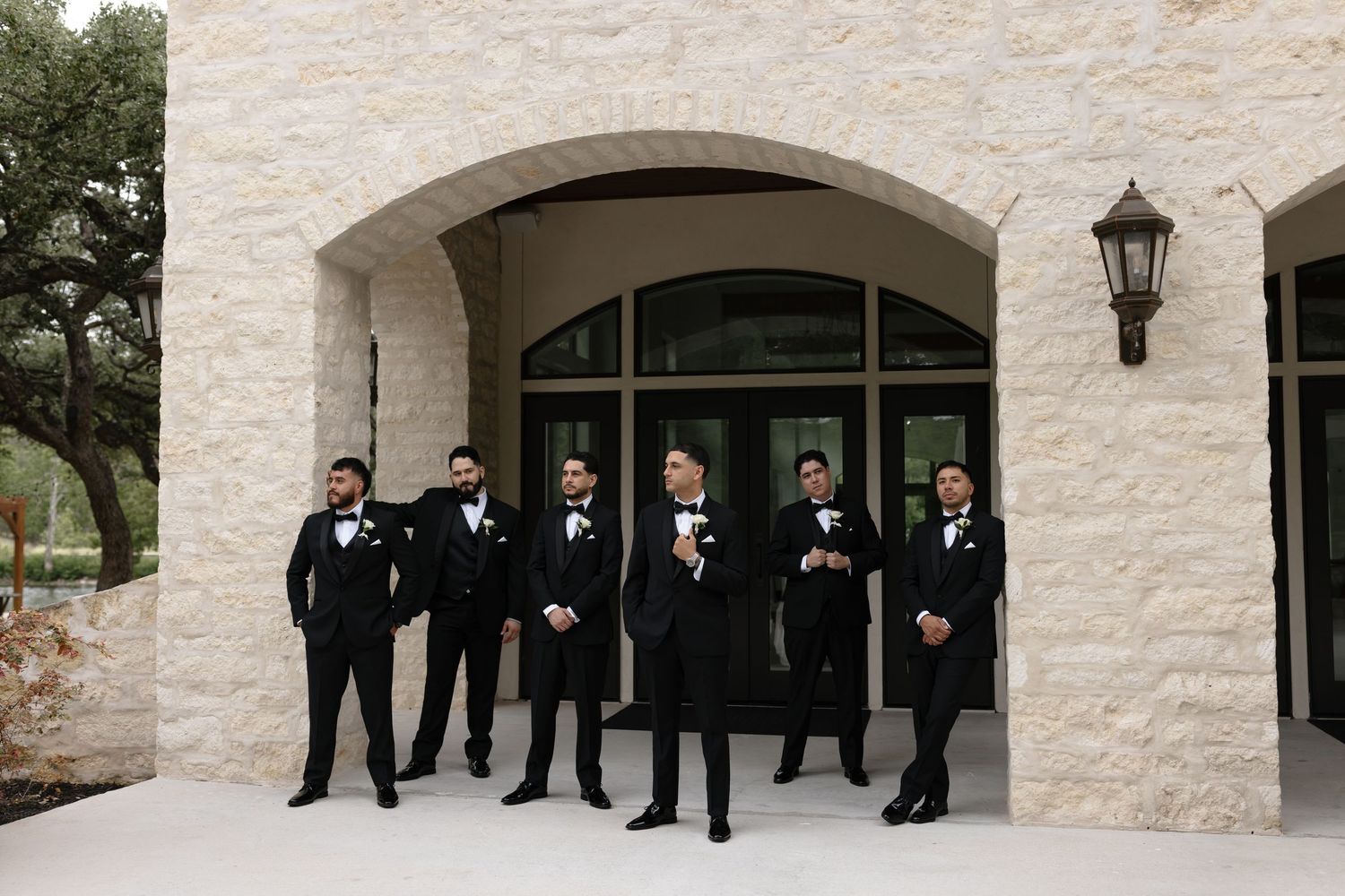 A group of groomsmen in black tuxedos stand together under a stone archway at an elegant wedding venue.