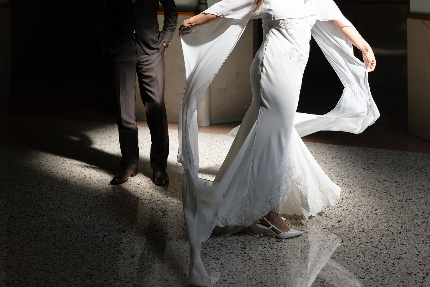 Bride in white mermaid wedding dress with flowing veil, assisted by groom in gray suit on marble floor.