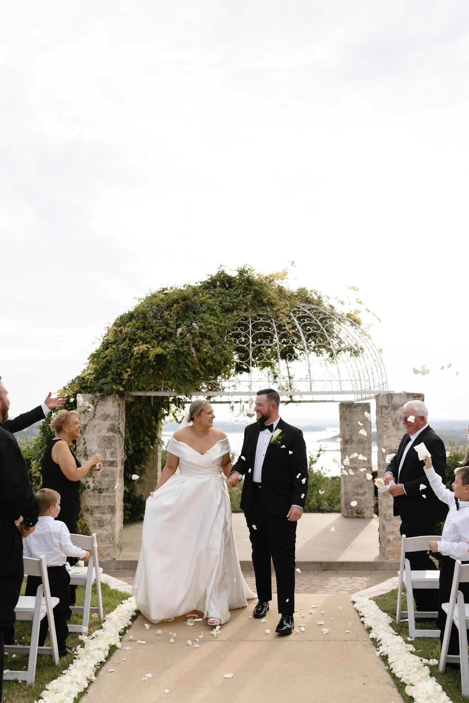 A couple walks down an outdoor aisle decorated with white petals under a vine-covered arch at a seaside wedding ceremony.