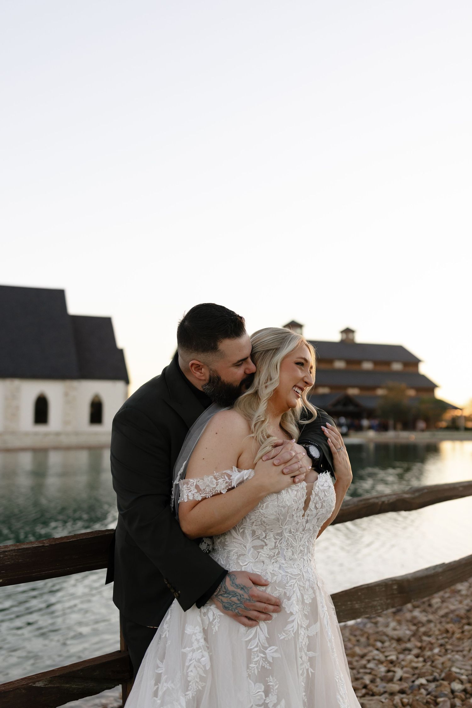 A couple in wedding attire shares an intimate moment by a waterfront at sunset.