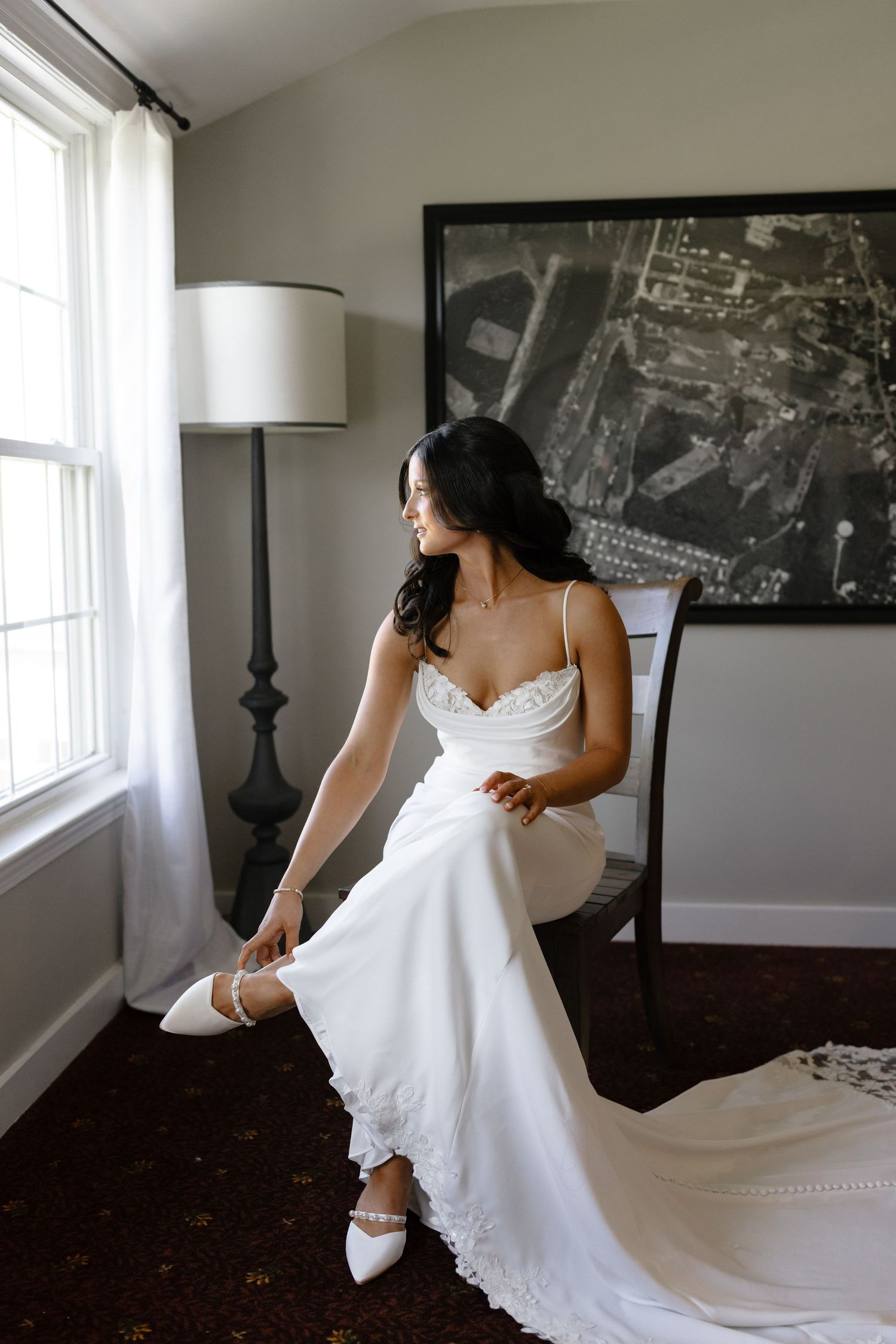 A bride in a flowing white wedding dress sits by a window adjusting her shoe, with a framed artwork on the wall behind her.