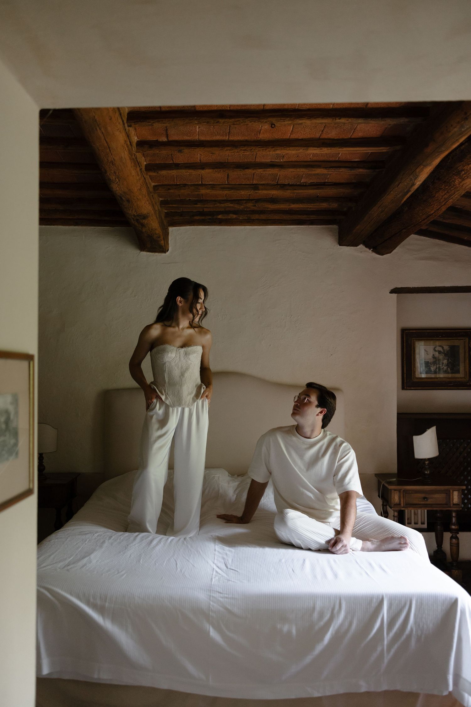 Couple in white outfits relaxing in rustic bedroom with wooden beam ceiling, woman standing on bed, man seated.