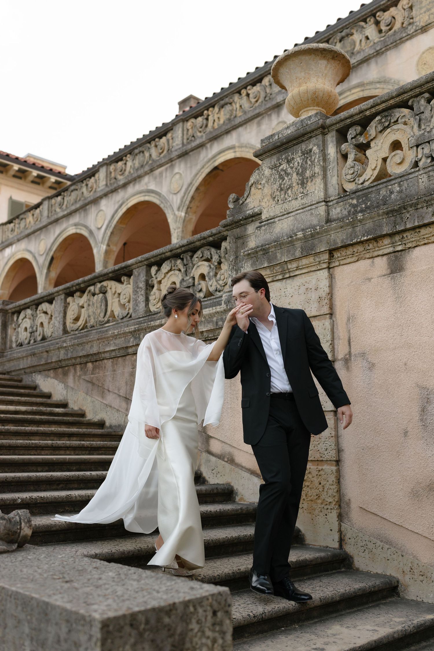 A couple in wedding attire descends ornate stone steps with arched architecture and decorative details in the background.