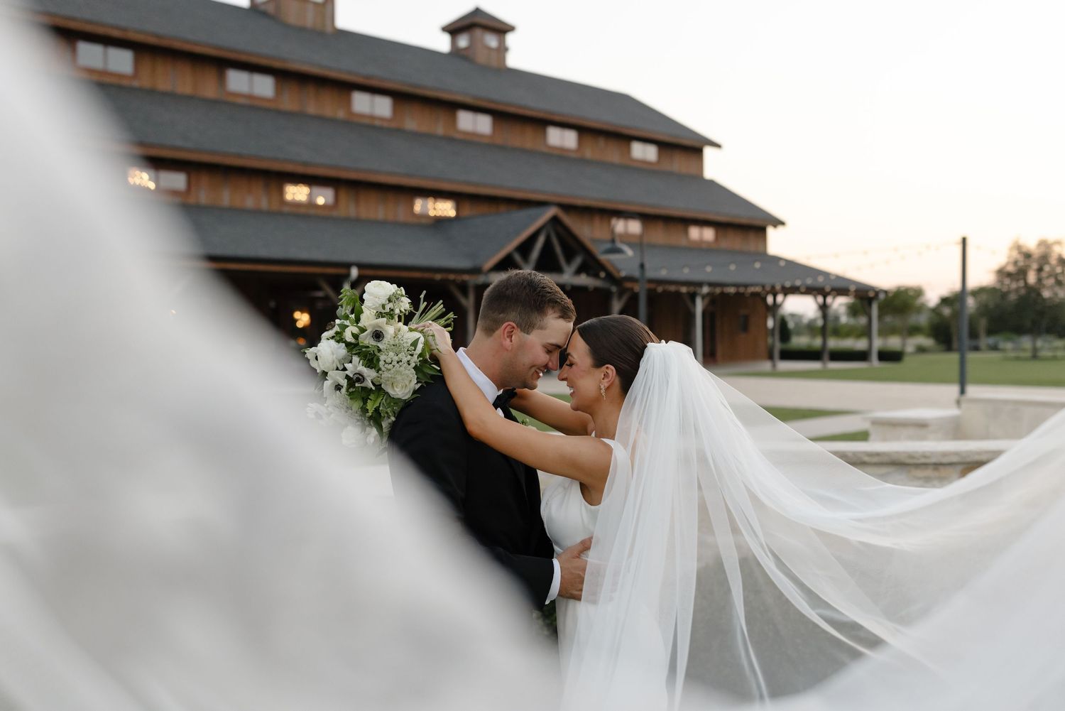 A flowing white wedding veil billows in the breeze against a rustic barn backdrop.