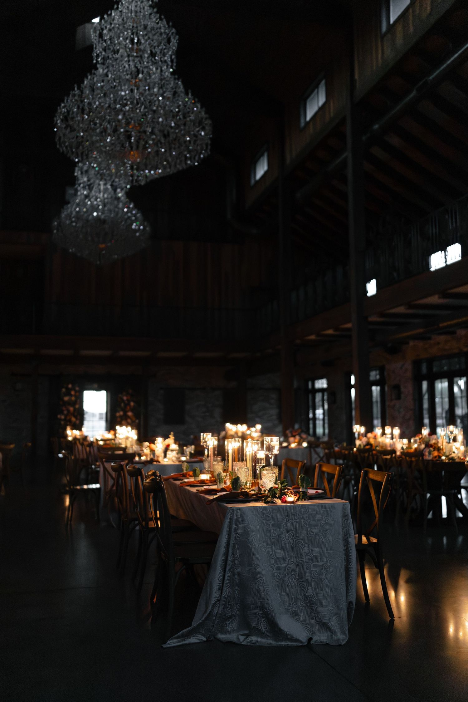 Elegant dark reception tables illuminated by crystal chandeliers create a dramatic atmosphere in an industrial venue.