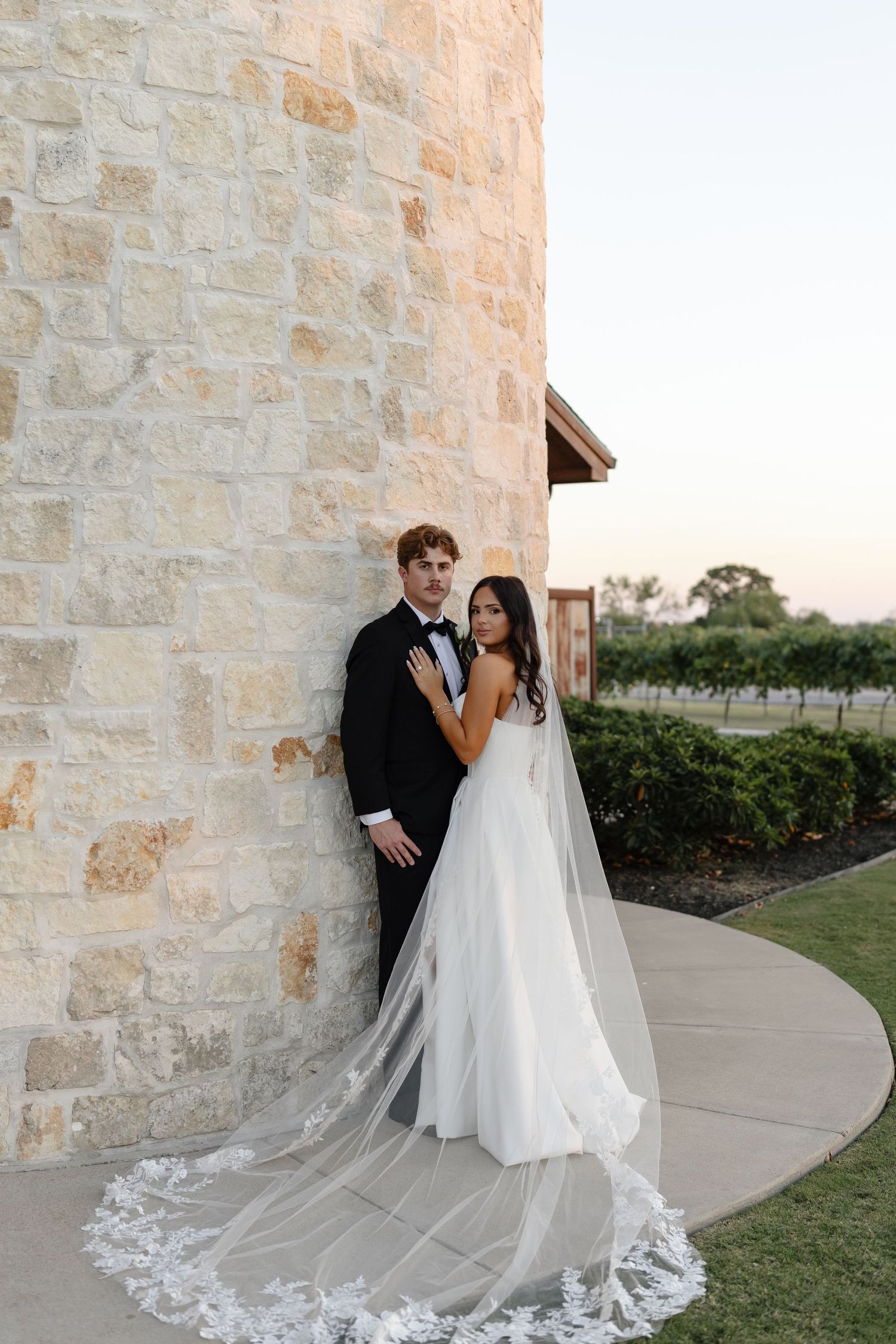 A romantic wedding couple poses against a stone wall exterior at sunset, with the bride's long lace veil trailing behind.