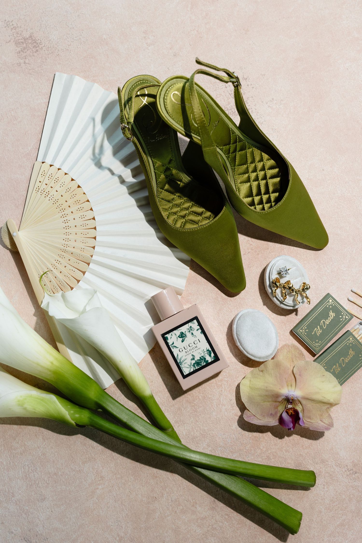 Flatlay of olive green slingback shoes with white paper fan, orchid flower, and beauty products on beige background.