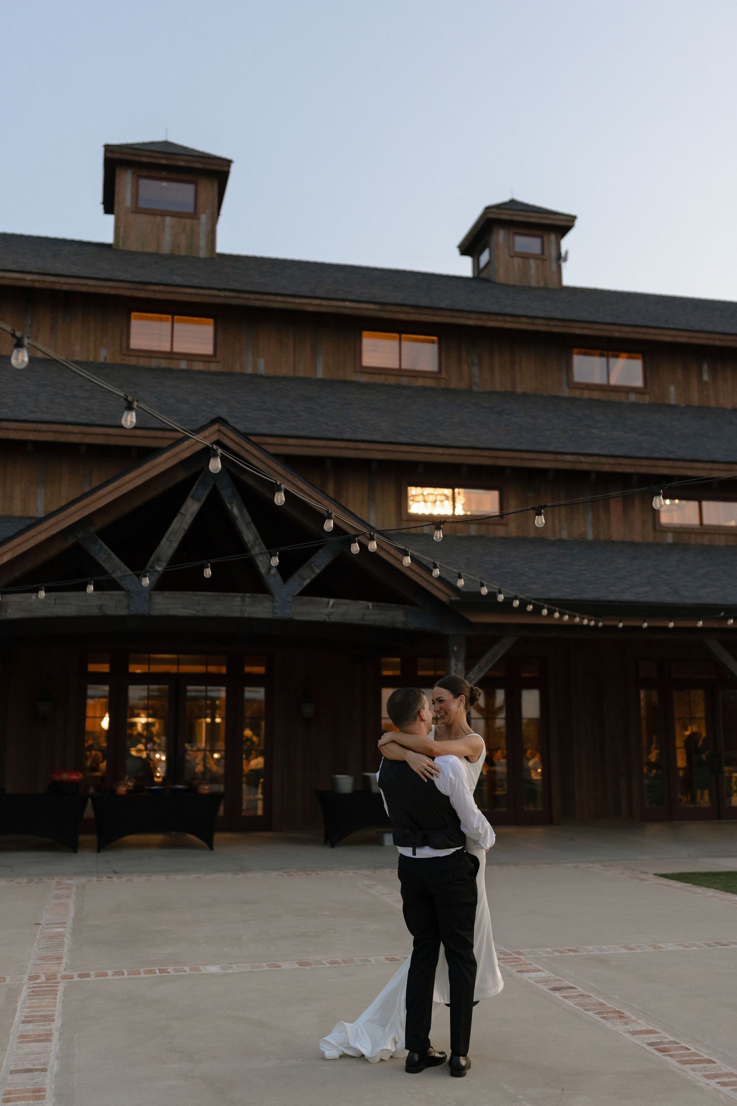 A couple shares a romantic moment outside a rustic wooden lodge with string lights at dusk.